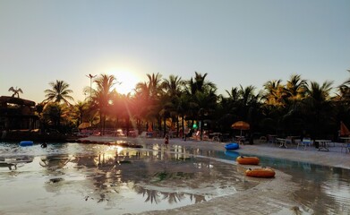 a view of sunset in a beach park, with palm trees, Floaters, beach chairs and sky