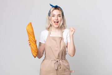 young pretty albino woman feeling shocked,laughing and celebrating success with a bread baguette