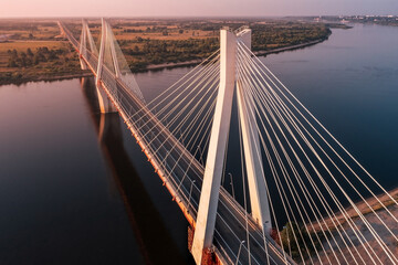 Diagonal aerial view of a white suspension bridge with three huge pillars above a river leading to a straight highway at sunrise in a rural area