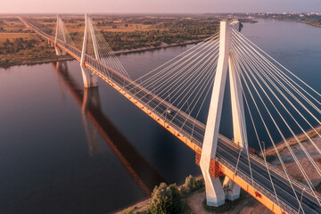 Diagonal aerial view of a white suspension bridge with three huge pillars above a river leading to a straight highway at sunrise in a rural area