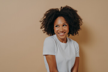 Cute african woman with toothy smile looking aside with shyness, isolated over beige background