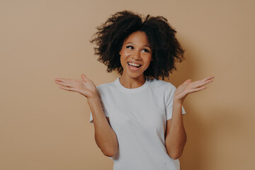 Young smiling mixed race woman raising hands with hesitation, isolated on beige background