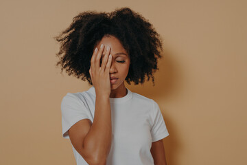 Dark skinned female covering face with hand and feeling anxiety or depression against beige wall