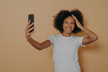 Happy charming dark skinned female making photo selfie on modern smartphone against beige wall