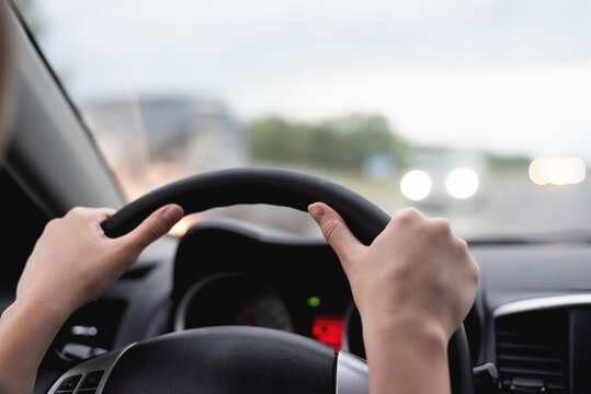 Female hands holds a car steering whell on the blurred cars background close up.