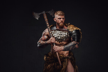 Fototapeta premium Brutal tattooed warrior wearing light armour and fur holding two-handed axe in dark studio