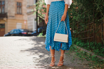 Summer street fashion details: elegant woman wearing trendy polka dot blue midi skirt, white strappy  sandals, holding classic leather bag, small handbag, posing in street. Copy, empty  space for text