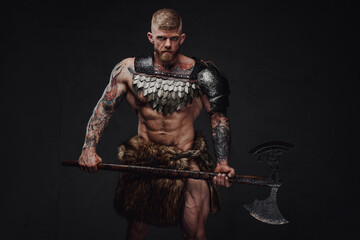 Fototapeta na wymiar Brutal tattooed warrior wearing light armour and fur holding two-handed axe in dark studio