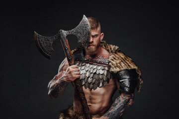 Brutal tattooed warrior wearing light armour and fur holding two-handed axe in dark studio