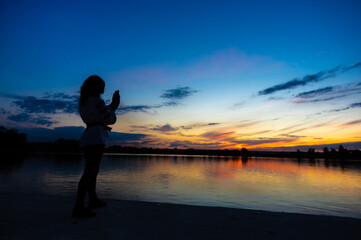 Fototapeta premium Silhouette of a girl with a phone on the background of sunset by the lake