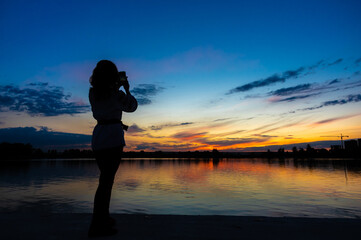 Silhouette of a girl with a phone on the background of sunset by the lake