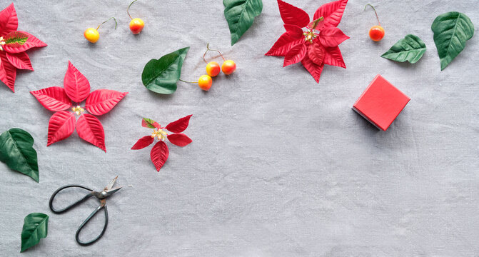 Christmas panoramic flat lay, banner on natural linen textile. Red poinsettia, Xmas decor, orange wild apples and red gift box. Scissors, red and dark green poinsettia leaves on beige textile.