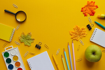 Back to school concept. Flat lay with different school supplies. Notebooks, pencils and pens, paper clips, paints, a magnifying glass, and a green apple on yellow background. Copy space. Top view