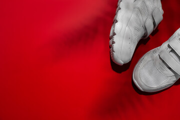 top view two white children's sneakers with velcro fasteners with shadows of palm or fern leaves isolated on a red saturated background.