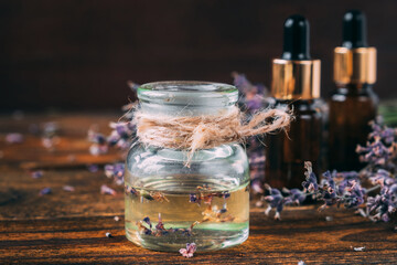 Lavender oil in a transparent jar tied with jute thread on a wooden table. In the background are oil bottles and a bouquet of lavender.