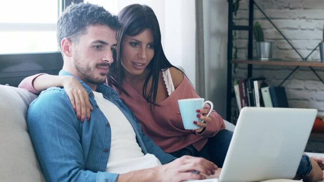 Video of happy beautiful couple using computer while sitting on the couch at home.