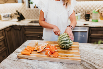 Woman slices watermelon, oranges, carrots, and ginger in her kitchen