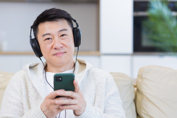 close up portrait of young asian man listening to music online in headphones using cellphone sitting on sofa at home