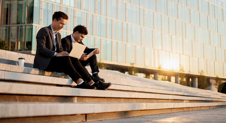 Successful colleagues work together. Businessmen young people work near the office in business suits holding gadgets in their hands. They are discussing a new startup project.