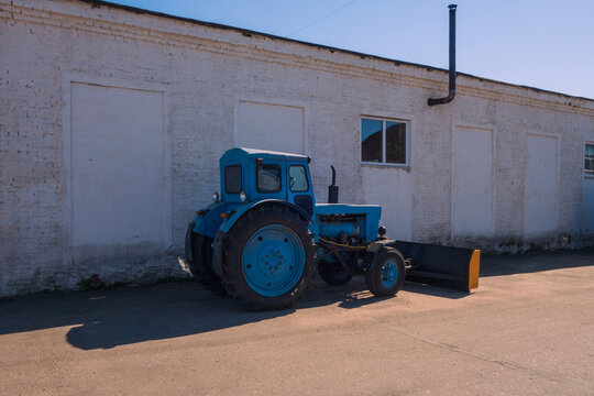 An old blue tractor stands on the asphalt near a white brick warehouse. Side view. Summer evening.