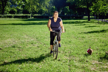 Young healthy woman rides bike in park with small dog running near. © grigvovan