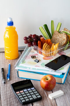 Close Up Of A Variety Of School Supplies With A Lunch Kit On Top And Bottle Of Juice To The Side, Against A Whiteboard.