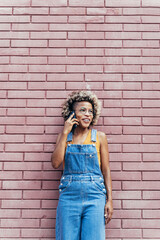 Stylish black African American Woman with blond hair and bib jeans on a phone call over a brick wall