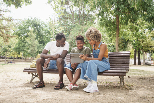 African American family having fun with tablet to watch online videos and social media. Young cool millennial parents and cute smiling kid sitting on bench in outdoor public park