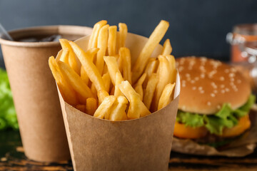 Paper box with tasty french fries, burger and cup of beverage on dark wooden background, closeup
