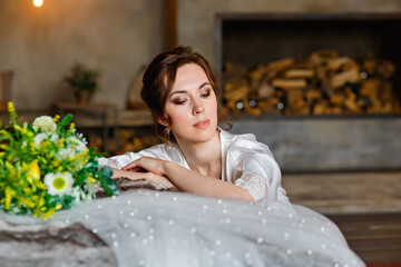 Naklejka premium A beautiful young brunette in a silk shirt sits next to the bed with a bouquet of flowers and a wedding veil. Bride's morning