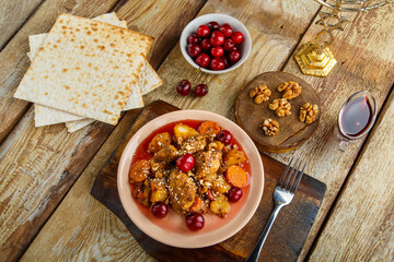 Jewish dish stewed potatoes with chicken in cherry sauce, decorated with cherries on a stand next to matzo and menorah.