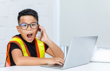 Young Asian boy wearing glasses and yellow safety vest as foreman woking on white office desk and exciting from receiving call contact by smartphone and surprise on interested info on laptop