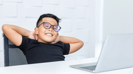 Fototapeta na wymiar Young Asian boy on black shirt happily resting on chair of white working desk of laptop in home office with hands behind head, smile, and eye close as satisfying and relieve on job success