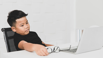 Poor Asian boy on black shirt seriously sitting on chair near white working desk of laptop in home...