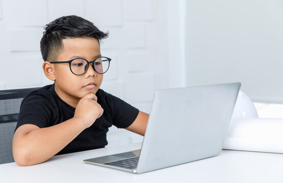 Smart Asian Boy Wearing Glasses And Black Shirt Sitting On Chair Of White Working Desk In Home Office, Seriously Catching Chin And Concentrate On Looking At Laptop To Consider Difficult Info