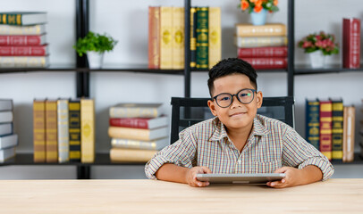 Fototapeta premium Smart Asian boy wearing glasses and grey checked shirt happily sitting in reading room and enjoy study of searching funny info by tablet on desk