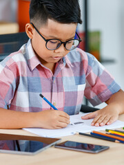 Naklejka na ściany i meble Smart Asian boy wearing glasses and grey checked shirt sitting on studying desk of tablet, mobile phone, and colorful pencils in reading room and concentrate on drawing on white paper