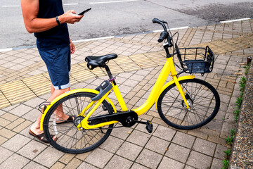Naklejka premium BIKE YELLOW and mobile phone. man using e vehicle rent service with smartphone in urban city street and park. Young man rent an eco transportation in summer