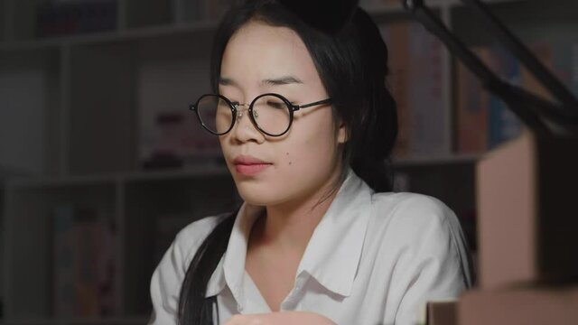 Close up tired young Asian woman wearing glasses, working hard till late night, checking stock and taking a note in working room at home, counting order, taking glasses off and getting headache
