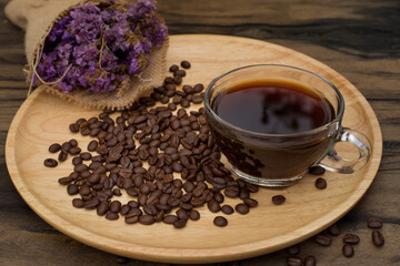 Black coffee in a cup and many coffee beans placed around on a wooden dish in a warm on wood background and bouquet of flowers purple color.