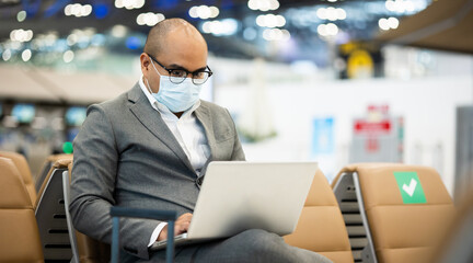 Businessman in airport terminal working with laptop . He wearing mask protection corona virus. He check in and waiting in passenger building.