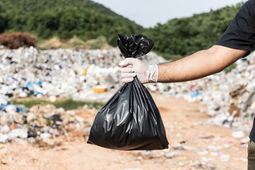 Fototapeta premium Hand holding a black trash bag in front of a large mountain and a pile of trash. Garbage plastics from industrial and community. World Environment Day.