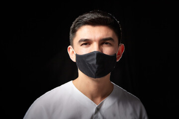 A young man who is a medical doctor in a medical mask a white uniform poses against a black background in the studio. Medic during the OKVED-19 pandemic