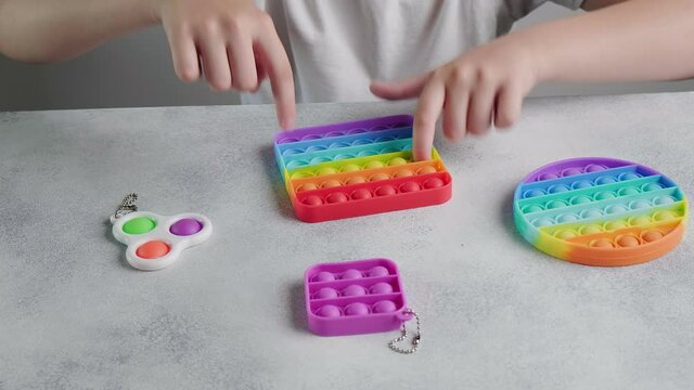 child playing pop it fidget toys, closeup on hands. unrecognizable little girl sits at table, pushes bubbles on colorful silicone poppit with fingers. close up shot of multicolor anti stress toys