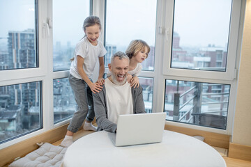 Gray-haired bearded man working on laptop while his kids interfering him