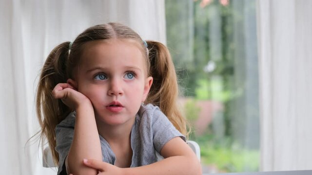 Bored preschooler girl blonde with long pigtails puts head on hands and looks straight sitting at home against window close view. Child holds head with hand and looks up and away dreaming