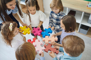 Team of little school children with teacher standing in circle and joining colorful jigsaw puzzle...