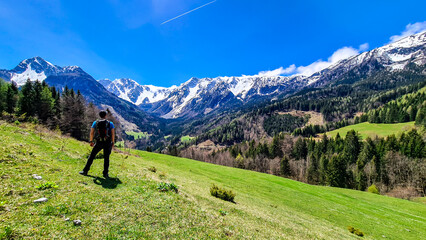 Fototapeta na wymiar A man with a big hiking backpack enjoying the panoramic view on Baeren Valley in Austrian Alps. The highest peaks are snow-capped. Lush green pasture. Clear and sunny day. High mountain chains.