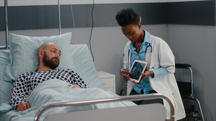 Fototapeta premium Afro american practitioner doctor analyzing bones radiography to sick man using tablet computer in hospital ward. Patient with nasal oxygen tube resting in bed during healthcare recovery