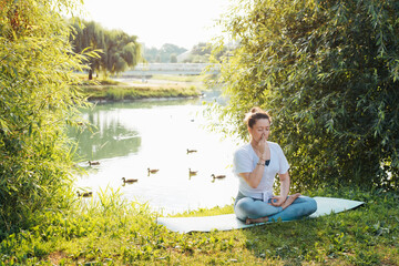 Young woman meditating by the lake, performing alternating nostril breathing, outdoors. Woman doing yoga asana nadi shodhana pranayama at sunrise in the park. Yoga and meditation in nature. - obrazy, fototapety, plakaty